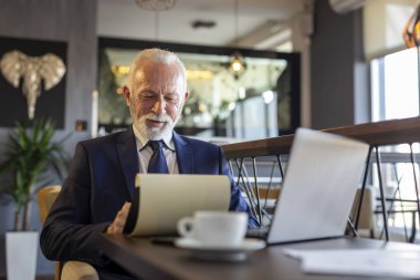 Restoran masasında oturan kıdemli işadamı dizüstü bilgisayarla çalışıyor, belgeleri ve sözleşmeleri analiz ediyor ve kahve içiyor.