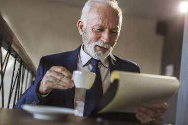 Restoran masasında oturan kıdemli işadamı, belgeleri ve sözleşmeleri analiz ediyor, notlar alıyor ve kahve içiyor.