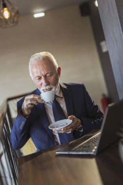 Restoran masasında oturan kıdemli işadamı, bir fincan kahve içiyor ve dizüstü bilgisayardan haber okuyor.