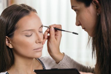 Makyaj stüdyosunda çalışan makyaj sanatçısı, kaş gölgesini kadın müşterinin kaşlarına uyguluyor.