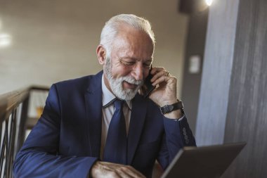 Modern bir ofiste çalışan kıdemli işadamı, dizüstü bilgisayarla uğraşırken telefon görüşmesi yapıyor.