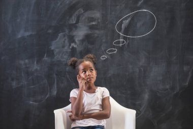 Güzel, melez bir ilkokul öğrencisi kafasının üstünde bulutlar olan düşünceli ve düşünceli bir kara tahtanın önünde oturuyor.