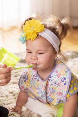 Beautiful little baby girl being fed by her mother, eating vegetable pap and smiling, smeared all over the face clipart