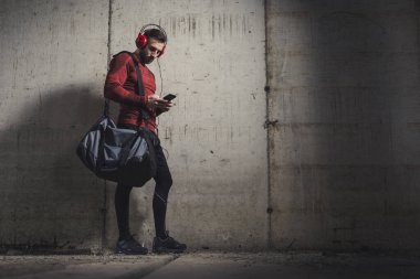 Spor kıyafetleri giyen, spor çantası taşıyan, müzik dinleyen ve spor yaptıktan sonra akıllı bir telefon kullanan kaslı bir estetik adam.