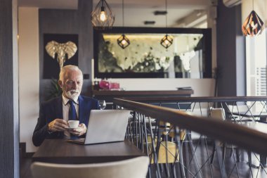 Restoran masasında oturan kıdemli işadamı, bir fincan kahve içiyor ve dizüstü bilgisayarında haberler okuyor.