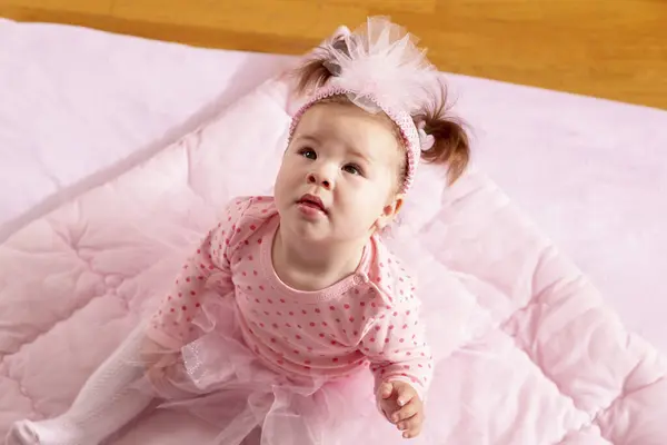 一个漂亮的小女孩穿着粉红的短裙坐在粉红的羽绒被上 面带微笑地抬起头 高视角 — 图库照片
