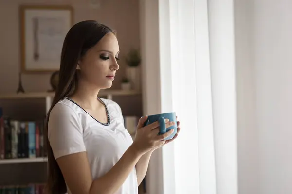 Beautiful young woman standing by the window and having her morning cup of coffee, relaxing and enjoying leisure time