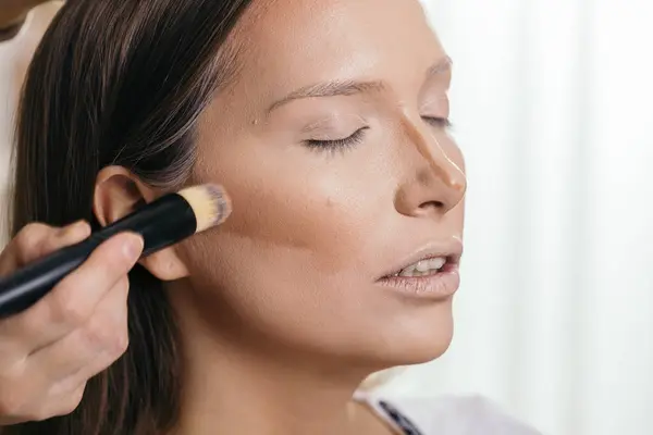 Make up artist applying liquid face powder foundation to a female client\'s face and blending contours