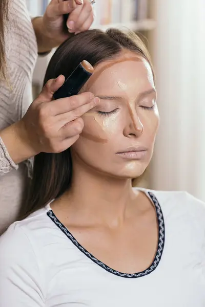 Make up artist applying face powder and corrector foundation to a female client\'s face, contouring the face