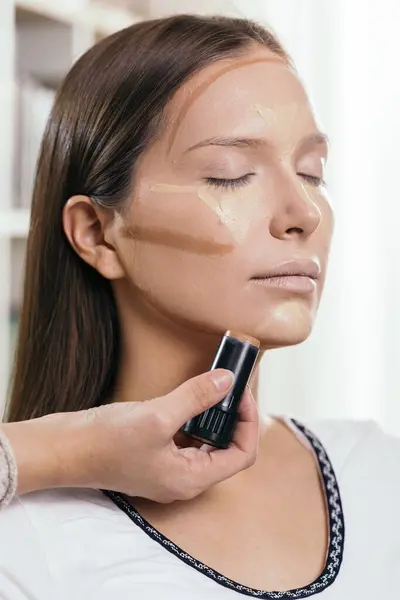 Make up artist applying face powder and corrector foundation to a female client's face, contouring the face