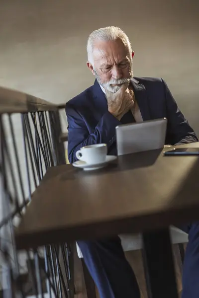 Senior businessman sitting at a restaurant table, drinking coffee and reading business news using a tablet computer, pensive and serious