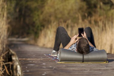 Göl kenarında uzanan bir adam, uzun bir doğa yürüyüşünden sonra, akıllı bir telefon kullanarak dinleniyor.