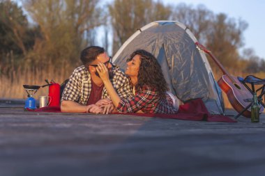Göl rıhtımında kamp yapan, çadır girişinde uzanan ve günbatımının tadını çıkaran bir çift.