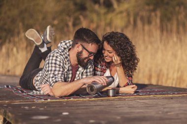 Genç çift göl rıhtımında piknik yapıyor, termostan kahve dolduruyor ve açık havada güneşli bir sonbahar gününün tadını çıkarıyor.