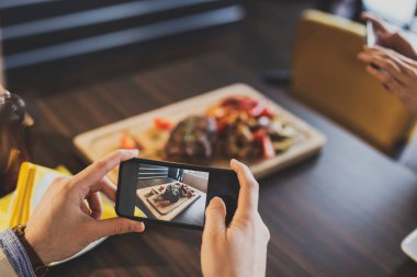 Restoran masasında oturan bir çift, öğle yemeğinden önce sipariş ettikleri yiyeceklerin fotoğraflarını çekiyor.