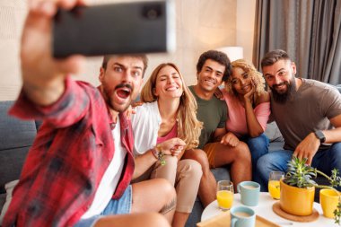 Bir grup neşeli genç arkadaş evde birlikte vakit geçirip, akıllı telefon kullanarak selfie çekiyorlar.