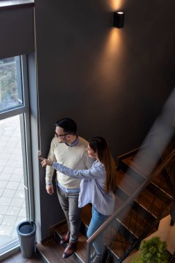 Sevgili bir çift, restoranın merdivenlerinden aşağı iniyor, el ele tutuşuyor, pencereden dışarı bakıyor ve dışarıdaki bir şeyi işaret ediyor.