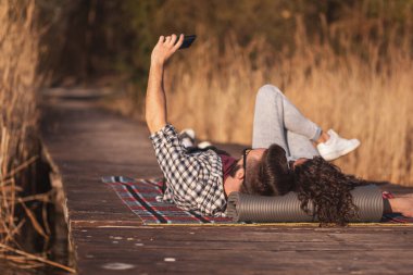 Güzel bir çift göl rıhtımında piknikte uzanmış güneşli bir sonbahar gününde selfie çekiyor ve eğleniyor.
