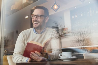Restoran masasında oturan, kahve içip kitap okuyan yakışıklı bir adamın portresi.