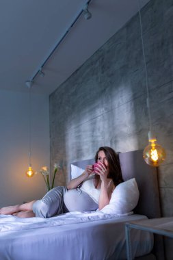 Gecelik giymiş güzel hamile kadın geceleri yatakta uzanıyor, çay içiyor ve evde dinleniyor.