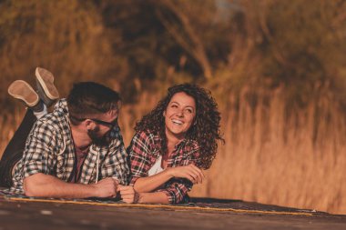 Birbirine aşık bir çift göl rıhtımında piknik battaniyesinde uzanıyor, eğleniyor ve güneşli bir sonbahar gününün tadını çıkarıyor.