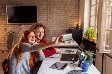 İki iş arkadaşı ofiste, akıllı bir telefonla selfie çekiyorlar; serbest çalışanlar birlikte çalışıyorlar ve tatilde eğleniyorlar.