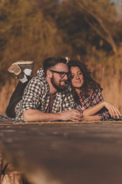 Birbirine aşık bir çift göl rıhtımında piknik battaniyesinde uzanıyor, eğleniyor ve güneşli bir sonbahar gününün tadını çıkarıyor.