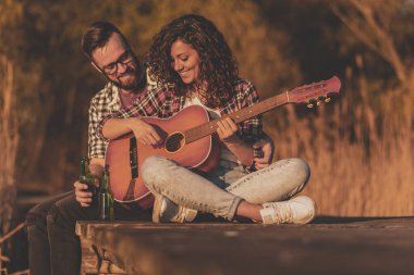 Göl rıhtımında oturan, gitar çalan, şarkı söyleyen, bira içen, doğanın tadını çıkaran ve dinlenen bir çift.