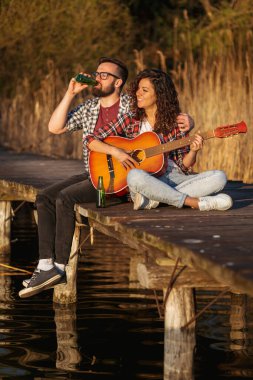 Göl rıhtımında oturan, gitar çalan, şarkı söyleyen, bira içen, doğanın tadını çıkaran ve dinlenen bir çift.