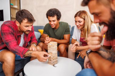 Bir grup neşeli genç arkadaş evde birlikte vakit geçiriyorlar, Jenga oynuyorlar, bir kuleden blokları sırayla kaldırıyorlar ve üstüne istifliyorlar.
