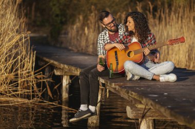 Göl rıhtımında oturan, gitar çalan, şarkı söyleyen, bira içen, doğanın tadını çıkaran ve dinlenen bir çift.