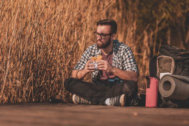 Yakışıklı erkek sırt çantalı, tahta göl rıhtımında oturuyor, yürüyüşe ara veriyor, doğada dinleniyor ve sandviç yiyor.