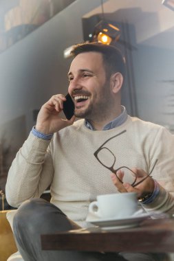 Restoran masasında oturan yakışıklı adam, telefonla konuşuyor ve kahve içiyor.
