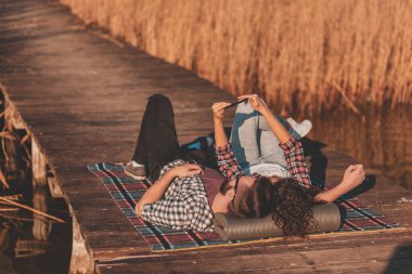 Güzel bir çift göl rıhtımında piknikte uzanmış güneşli bir sonbahar gününde selfie çekiyor ve eğleniyor.