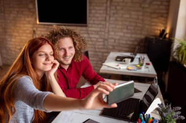 İki iş arkadaşı ofiste, akıllı bir telefonla selfie çekiyorlar; serbest çalışanlar birlikte çalışıyorlar ve tatilde eğleniyorlar.