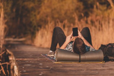 Göl rıhtımında uzanan adam, uzun bir doğa yürüyüşü gününden sonra dinleniyor, akıllı bir telefon kullanıyor, açık havada çalışan özgür bir erkek.