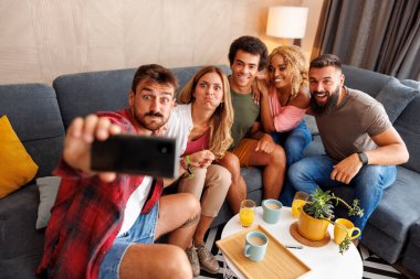 Bir grup neşeli genç arkadaş evde birlikte vakit geçirip, akıllı telefon kullanarak selfie çekiyorlar.