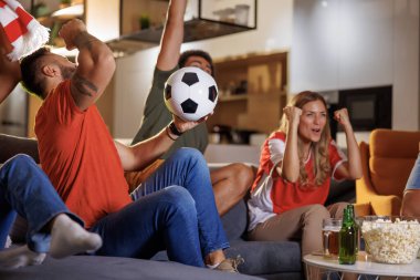 Bir grup futbol taraftarı televizyonda maç izlerken tezahürat yapıyor, takımlarının gol atmasını ve maçı kazanmasını kutluyorlar. Futbol topuna odaklan.