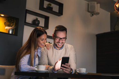 Restoran masasında oturan, kahve içen ve akıllı telefon kullanan genç ve güzel bir çift.