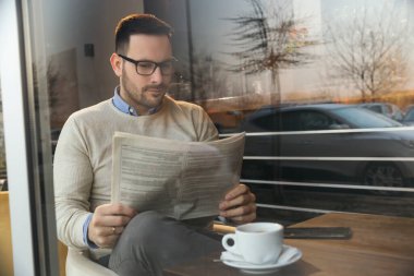 Yakışıklı bir adam restoran masasında oturuyor, gazete okuyor ve kahve içiyor.