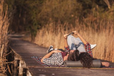 Güzel bir çift göl rıhtımında piknikte uzanmış güneşli bir sonbahar gününde selfie çekiyor ve eğleniyor.