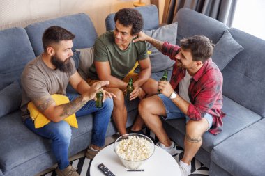 Bir grup erkek arkadaşın evde birlikte vakit geçirip, televizyon izleyerek, patlamış mısır yiyerek ve bira içerek eğlendiği geniş açılı bir manzara.