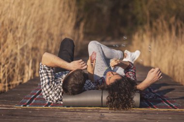 Birbirine aşık bir çift, göl rıhtımında piknik battaniyesinde uzanmış, sabun köpüğü yaparken eğleniyor ve doğada güneşli bir sonbahar gününün tadını çıkarıyor.