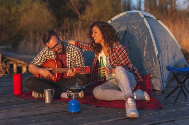 Güneşli bir sonbahar gününde göl kenarında kamp yapan çift gitar çalıyor, bira içiyor ve dışarıda eğleniyor.