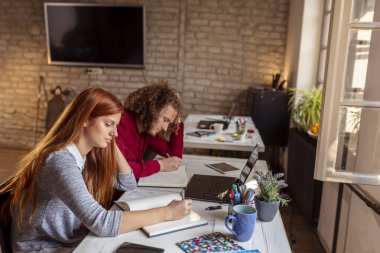 Modern bir ofiste çalışan iş arkadaşları, sabah brifingleri sırasında planlamacılarda notlar alıyorlar; çalışma alanında çalışan serbest çalışanlar