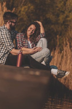 Genç çift göl rıhtımında oturmuş, doğada güneşli bir sonbahar gününün tadını çıkarıyorlar.