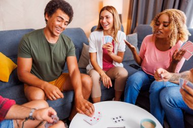 Neşeli genç arkadaşlar evde birlikte vakit geçiriyorlar, kağıt oynayarak eğleniyorlar.