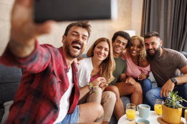 Bir grup neşeli genç arkadaş evde birlikte vakit geçirip, akıllı telefon kullanarak selfie çekiyorlar.