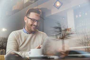 Yakışıklı genç adam restoran masasında oturuyor, kahve içiyor ve tablet bilgisayar kullanıyor.