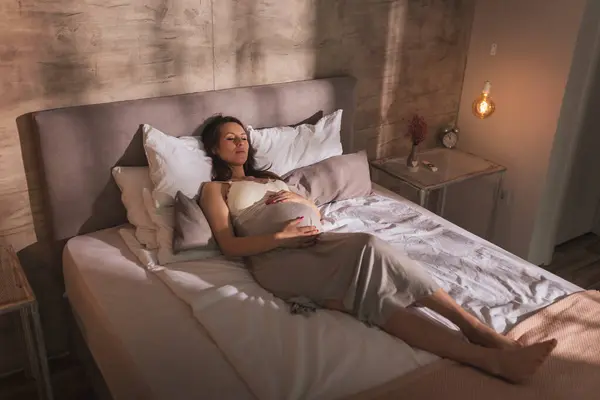 ナイトガウンを着た美しい妊婦の肖像画 ベッドに横たわり 腹を抱えて寝ている — ストック写真
